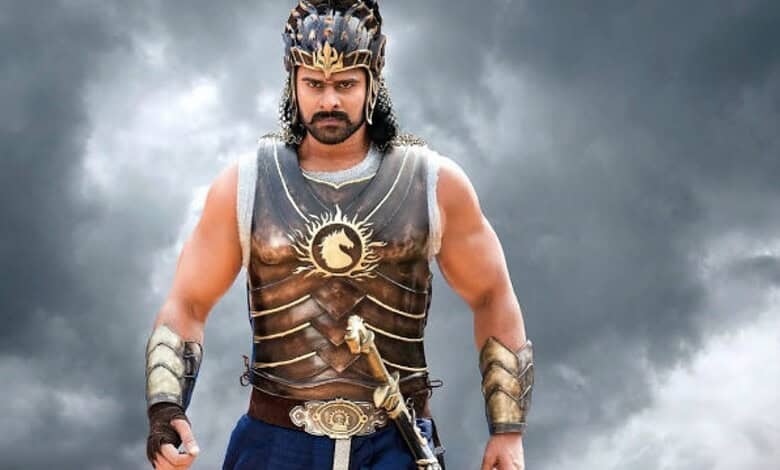 Prabhas as Baahubali wearing armor and crown, standing confidently against stormy sky.