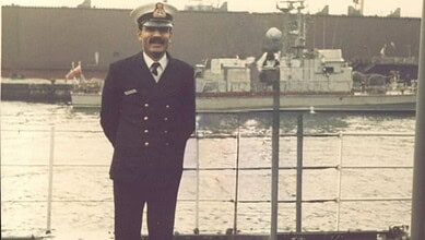 Indian Navy officer in uniform standing by the water with ships in the background.