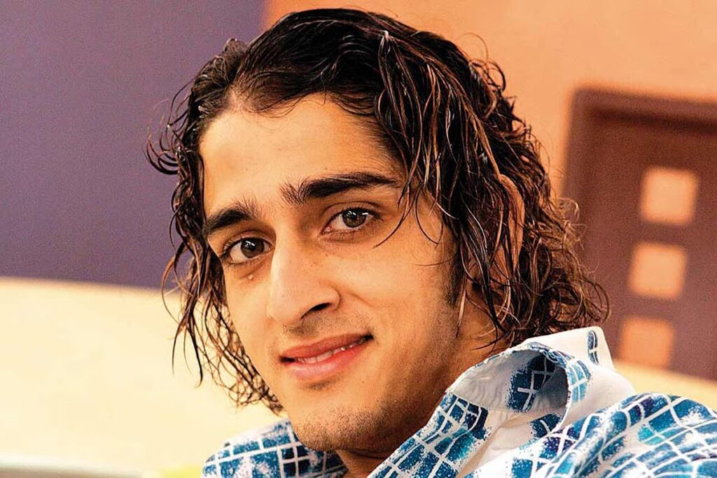 Young man with curly hair and a blue patterned shirt smiling at the camera.