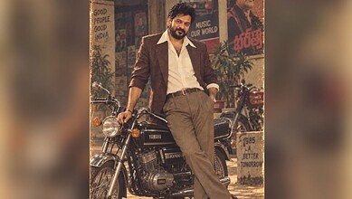 Revanth Reddy in 1980s style outfit with vintage motorcycle in a retro-themed setting.