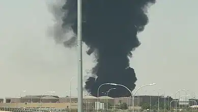 Thick black smoke rising from a fire near Riyadh airport, with clear sky and streetlights in the scene.