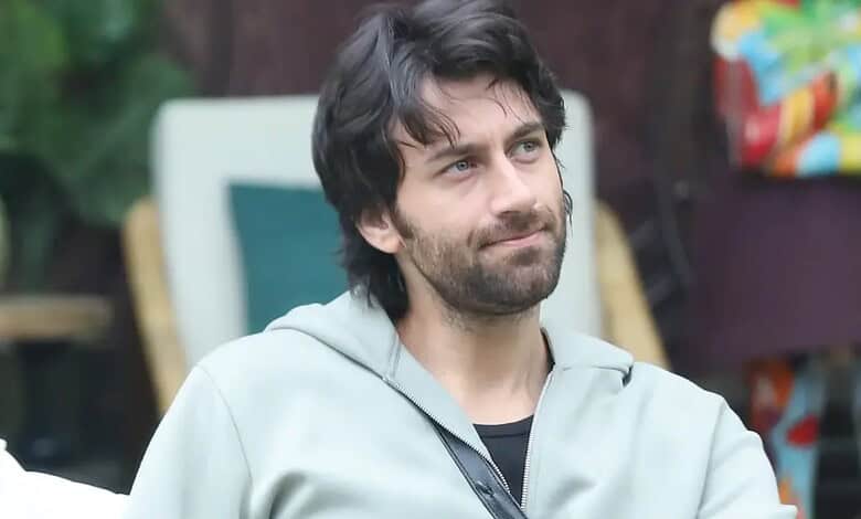 Man with dark hair and beard wearing a light grey hoodie outdoors.