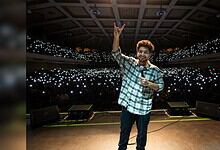 Samay Raina performing on stage with a large audience and bright lights in the background.