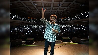 Samay Raina performing on stage with a large audience and bright lights in the background.