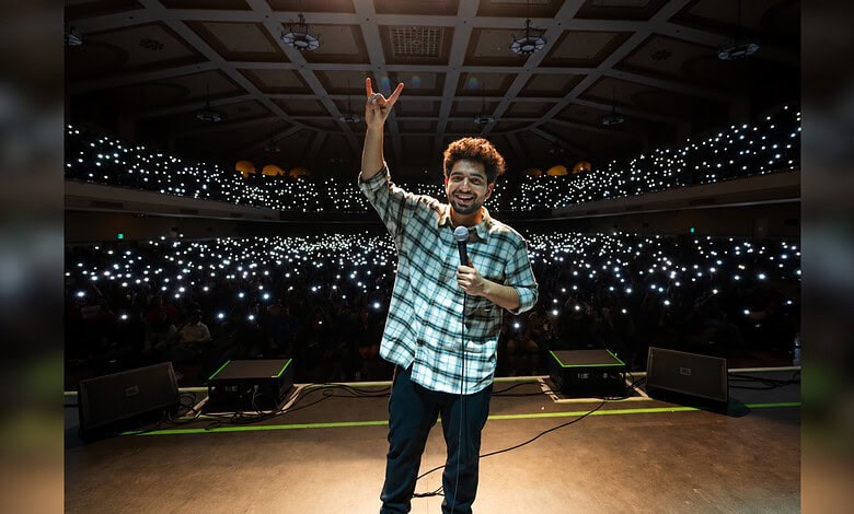 Samay Raina performing on stage with a large audience and bright lights in the background.