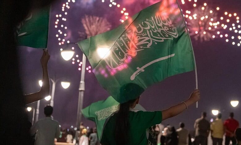 People celebrating Saudi Arabia National Day with fireworks and flags at night.