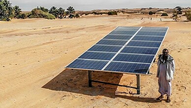 Man standing beside solar panel in arid Sudan desert with sparse vegetation.