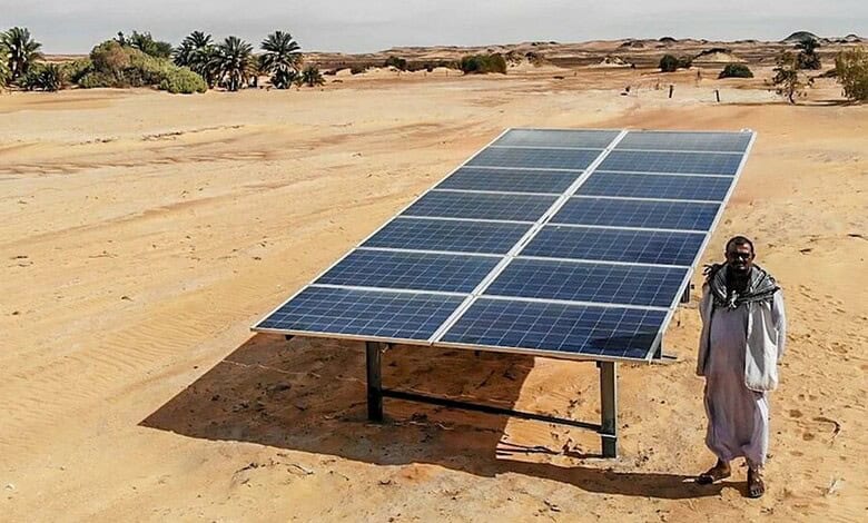 Man standing beside solar panel in arid Sudan desert with sparse vegetation.