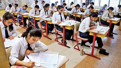 Students in white uniforms taking Class 12 exams in a well-lit classroom.