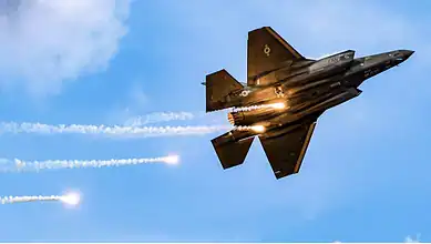 F-35 fighter jet flying at high speed with afterburners on against blue sky.