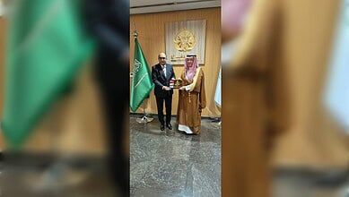 Two men exchanging awards at a formal event with flags and emblem in the background.