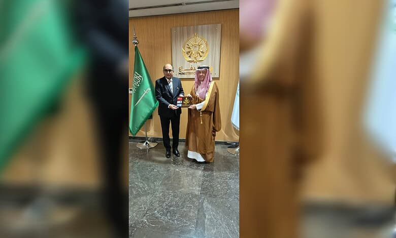 Two men exchanging awards at a formal event with flags and emblem in the background.