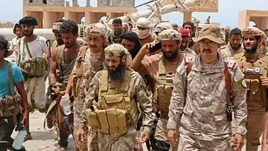 Yemeni soldiers in combat gear walking through a desert area with buildings in the background.