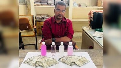Man sitting at a desk with cash and bottles of pink and clear liquids in front of him.