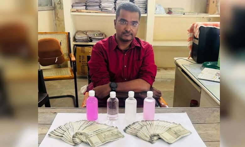 Man sitting at a desk with cash and bottles of pink and clear liquids in front of him.