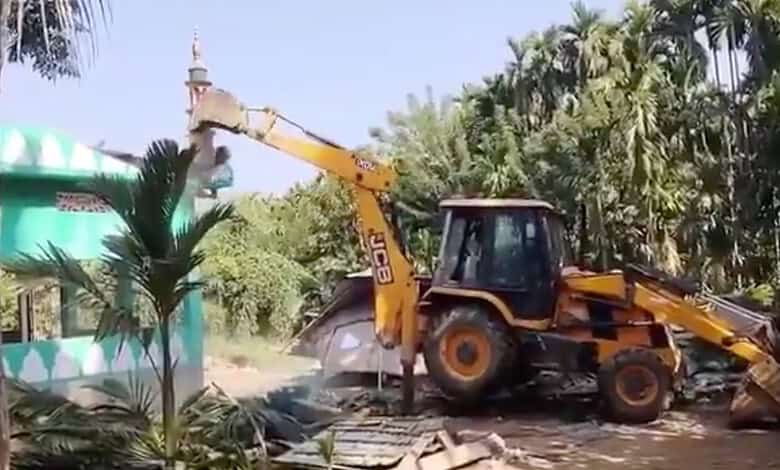 Excavator demolishing structures amidst greenery in Assam.