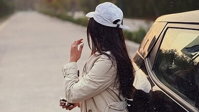 Woman with long dark hair wearing a white cap and beige jacket standing near a car on a road.