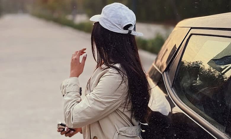 Woman with long dark hair wearing a white cap and beige jacket standing near a car on a road.