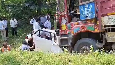 A large truck crashes into a white car in a grassy, wooded area with onlookers nearby.