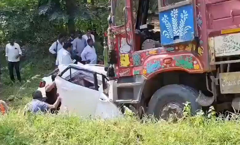 A large truck crashes into a white car in a grassy, wooded area with onlookers nearby.