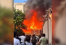 Large fire with thick black smoke near reality show sets, crowd watching from a distance.