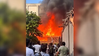Large fire with thick black smoke near reality show sets, crowd watching from a distance.
