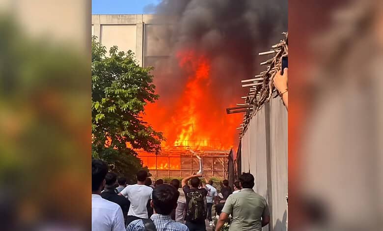 Large fire with thick black smoke near reality show sets, crowd watching from a distance.