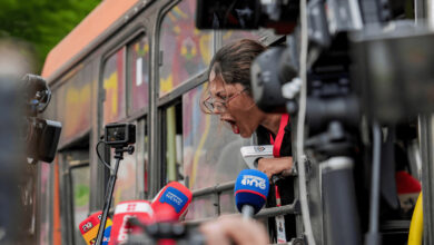 Journalist shouting from a bus window during the protest, surrounded by microphones and cameras.