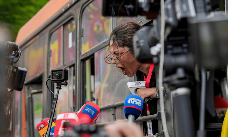 Journalist shouting from a bus window during the protest, surrounded by microphones and cameras.