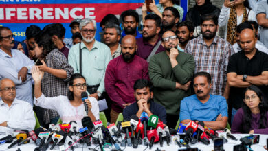 Group of people at a press conference with microphones and cameras in front.