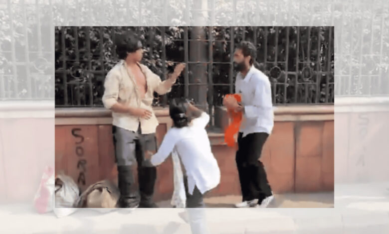 Two men and a woman engaged in a heated argument during a protest in Delhi, with a metal fence in the背景.