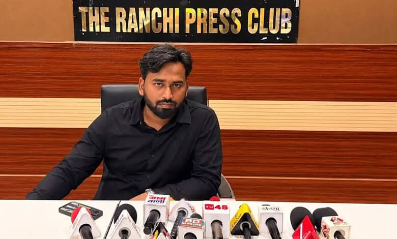 Man in black shirt at press conference with microphones in front, 'The Ranchi Press Club' sign behind.