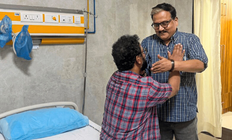 Two men in a hospital room, one sitting on the bed and the other standing, engaging in a conversation.