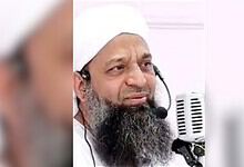 Close-up of a Muslim preacher with a white turban and beard speaking into a microphone.