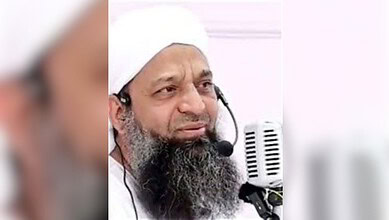 Close-up of a Muslim preacher with a white turban and beard speaking into a microphone.