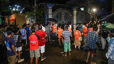 Crowd gathered outside a gated area during night, some with umbrellas, in Kolkata.