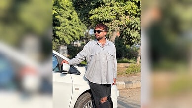 Young man with sunglasses and ripped jeans standing beside a white car in a park.