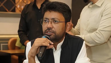 Man speaking into a microphone at a formal event, wearing glasses and a black vest.