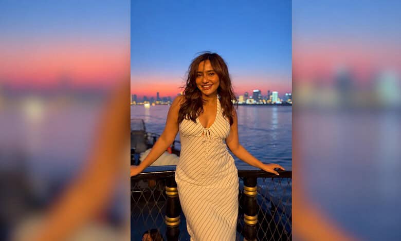 Neha Sharma smiling by the river during sunset with city skyline in background.
