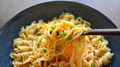 Chopsticks lifting a portion of cooked instant noodles with green onions.