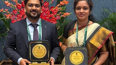 Two Telangana officials holding awards at a formal event with vibrant red and green plants in the背景.