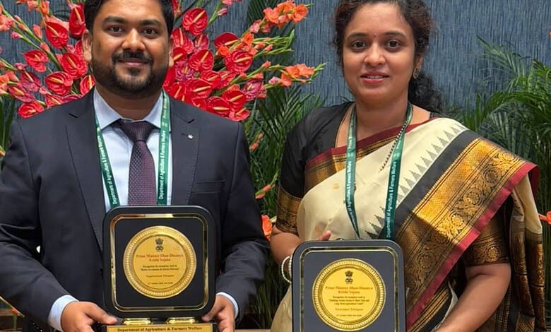Two Telangana officials holding awards at a formal event with vibrant red and green plants in the背景.