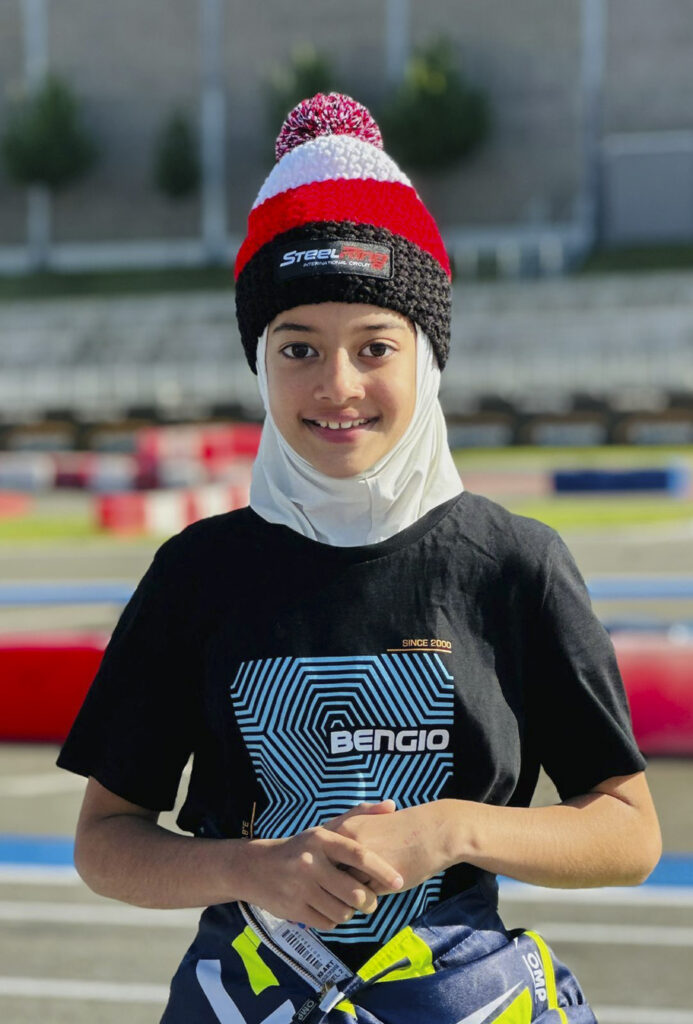 Indian girl Atiqa Mir in racing gear at karting track, smiling for the camera.