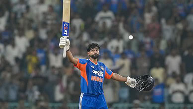 Indian cricket player in blue jersey holding bat and helmet on the field.