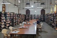 Students studying in a library with tall bookshelves and large arched windows.