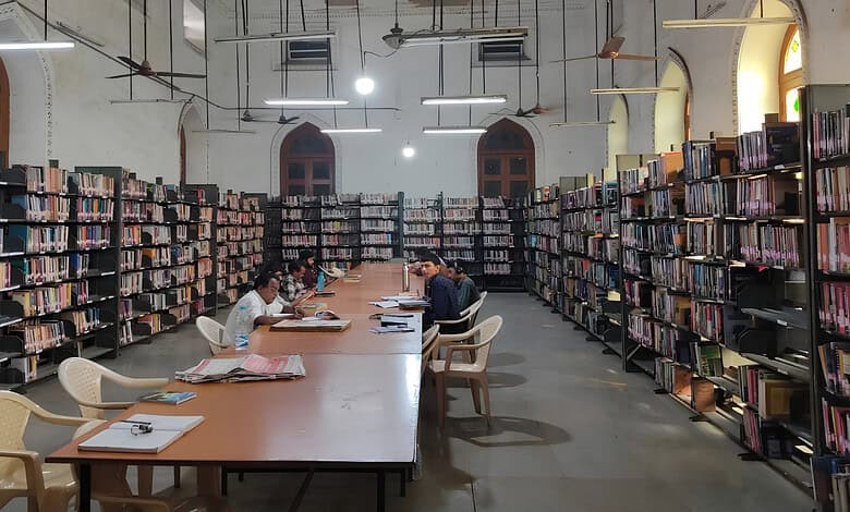 Students studying in a library with tall bookshelves and large arched windows.