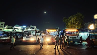 Nighttime scene at a bus station with multiple buses and people under a dark sky.