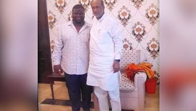 Two men standing together in a room with floral wallpaper and a sofa.