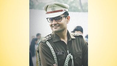 Smiling IPS officer in uniform with glasses and cap, outdoors with blurred crowd background.