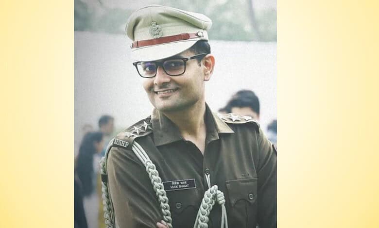 Smiling IPS officer in uniform with glasses and cap, outdoors with blurred crowd background.